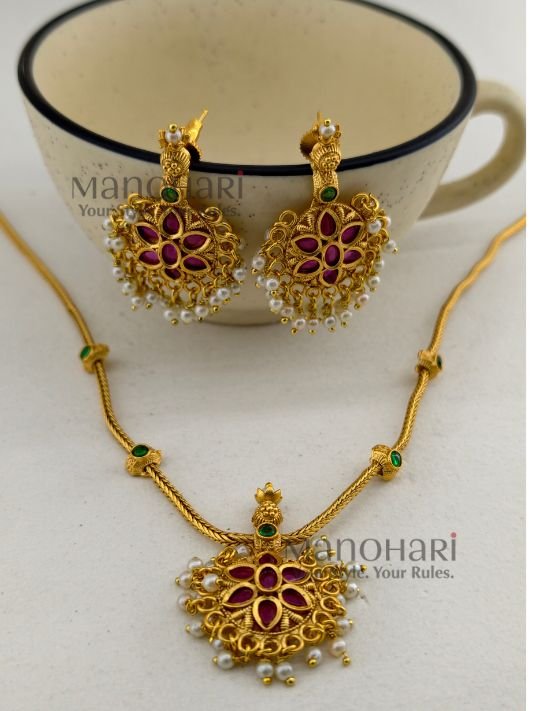 MN-NK-KMP-MAK-01_2 **The Meher Antique Kemp Necklace Set is a timeless celebration of South Indian heritage, crafted from **premium brass** with a luxurious **matte gold-plated finish**. Embellished with vibrant **traditional Kemp stones**, this elegant necklace showcases intricate temple-inspired craftsmanship that radiates regal charm. The **21 cm necklace** features an **adjustable extension chain** for a comfortable fit, while the matching **4.5 cm earrings** complete the coordinated look. Perfectly paired with **Kerala Kasavu sarees, Kanchipuram silk sarees, lehengas, Anarkalis, and other traditional attire**, this exquisite jewellery set is ideal for **Onam, weddings, temple visits, engagements, receptions, festivals, and bridal celebrations**. Exclusively curated by **Manohari Style**, the **Meher Antique Kemp Necklace Set** combines heritage artistry, premium craftsmanship, and timeless elegance, making it a treasured addition to every woman's ethnic jewellery collection.**